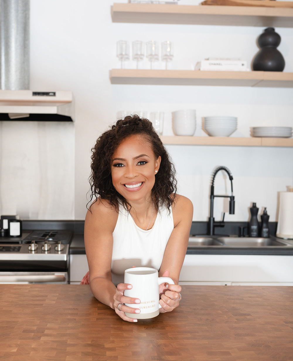 Gabriella leaning on kitchen island while holding coffee mug.