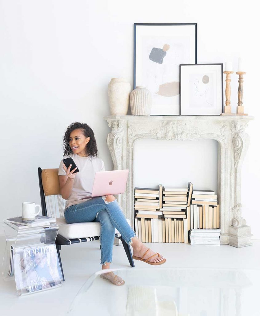 Gabriella sitting in front of books and fireplace with laptop on lap and phone in hands.