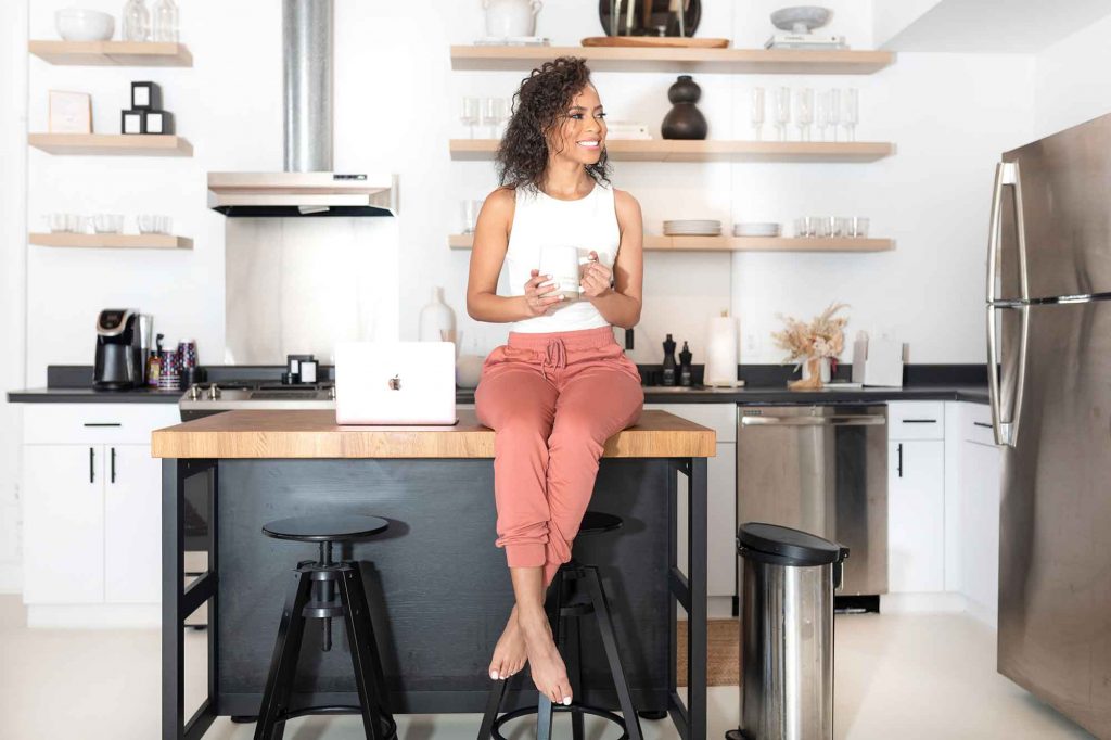 Gabriella sitting on kitchen table with coffee mug in hand and laptop beside her.